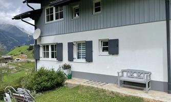 A charming house with gray shutters and a green garden. In the background, gentle hills and mountains can be seen.