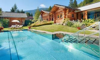An elegant wooden house with a beautiful terrace and a pool area. Surrounded by a green garden and mountains in the background.