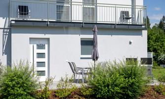 A modern house with a balcony and a small outdoor seating area. Surrounded by green plants and a clear sky.