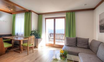 A bright living room with a cozy gray sofa and a dining table. Large windows with green curtains offer a beautiful view outside.