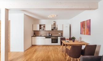 A modern kitchen with wooden flooring and a dining area. Colored pictures hang on the wall, adding color to the room.