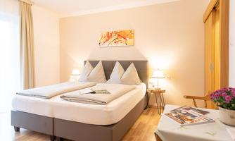 A bright and modern bedroom with a double bed and colorful cushions. Next to the bed are nightstands, and there is a small table with a flower.