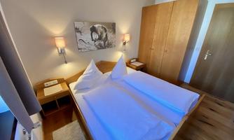 A bright bedroom with a double bed and two pillows. The room features wooden wardrobes and soft lighting.