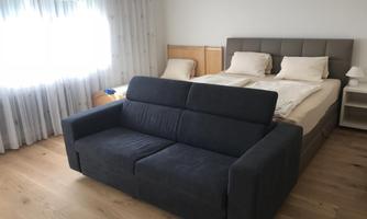 A bright bedroom with a large double bed and a gray sofa. The room has large windows with curtains and a friendly wooden floor.