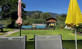 A quiet garden with sun loungers and umbrellas. In the background, there is a pond and a wooden hut.