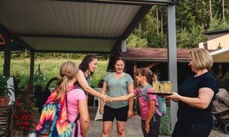 A group of women and girls greets each other happily outdoors. One woman serves drinks while the others chat and laugh.