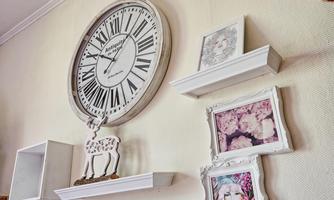 A large wall clock with Roman numerals hangs on a wall. Below it are white shelves with various pictures and a small decorative object.