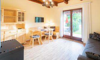 A cozy living space with a sofa, a dining table, and a television. Large windows offer a view of a garden area.