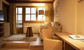 A cozy living room with wooden details and large windows. There is a sofa, a table, and a desk area with warm light.