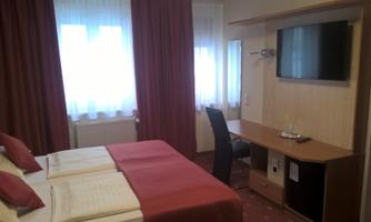 A cozy hotel room with two single beds and a television. Large windows provide plenty of daylight.