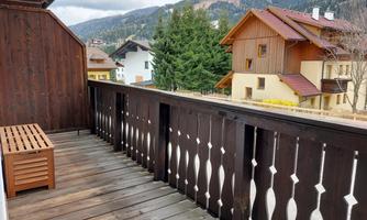 A wooden balcony with a cozy seating area. In the background, green trees and houses, as well as mountains, are visible.