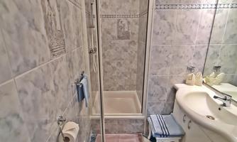 A small bathroom with a shower, sink, and toilet area. The walls are tiled in light colors, and the floor has a patterned design.