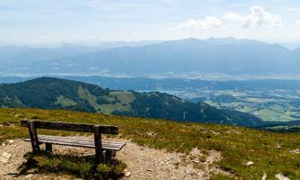 A wooden bench sits on a mountain overlooking the valley. The landscape is green and the mountains in the background are in soft colors.
