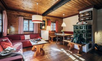 A cozy living room with a wooden ceiling and floor. There is a red couch, a table, and a traditional stove.