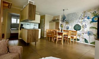 A modern living room with an open kitchen and a dining table for six people. The wall is decorated with colorful floral motifs.