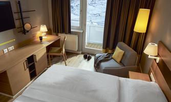 A cozy hotel room with a large bed, a desk, and an armchair. The windows let in plenty of daylight and offer a view outside.