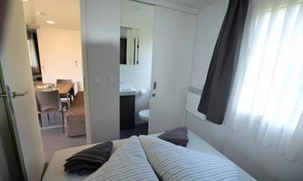 A modern bedroom with a cozy bed and striped pillows. In the background, a bright living and dining room is visible.