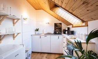 A modern kitchen under a sloping roof with wooden beams. The decor is bright and welcoming, with plants and kitchen utensils.