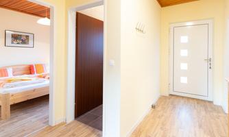 A bright hallway with a white entrance door and wooden floor. On the left side is a room with a bed and colorful bedding.