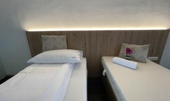 A modern bedroom with two single beds and soft lighting. The pillows are decorative and a flower is placed on one of the beds.
