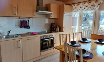 A modern kitchen with wooden furniture and a large window front. There is a dining table set with dishes and a well-equipped cooking area.