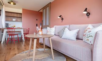 A cozy living room with a light pink sofa and affectionate cushions. On the table are wine and fresh flowers, while red chairs and a warm wooden floor create an inviting atmosphere.