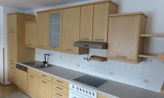 A modern kitchen with light wood cabinets and a simple countertop. The equipment includes a stove, a sink, and open storage.