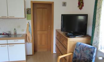 A small apartment with a kitchenette, a TV, and a wooden cupboard. Next to it stands a blue armchair and a table with a chair.