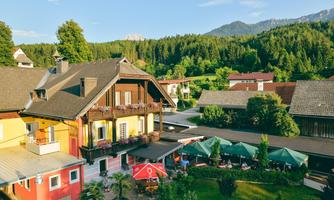 A picturesque country house surrounded by green forests and mountains. In the foreground, there is a cozy garden with sun umbrellas and seating areas.
