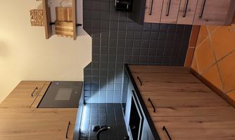 A modern kitchen with wooden cabinets and dark tiles. The room is well-lit and contains a sink as well as kitchen appliances.