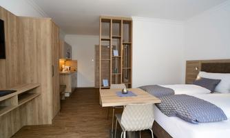 A modern room with a double bed, a small dining table, and a kitchenette. The decor is bright and friendly, with wooden surfaces and minimalist design.