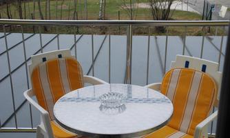 A balcony with two white chairs and a round table. The table has an ashtray and overlooks a green meadow.