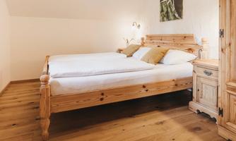A cozy bedroom with a wooden bed frame and bright pillows. The floor is made of wood, and there is a rustic wardrobe and soft lighting.