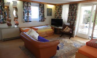 A cozy living room with a brown couch, a television, and a rug. The walls are decorated with colorful curtains and there is plenty of daylight.