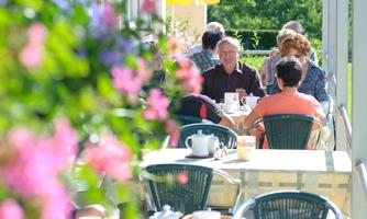 A cozy outdoor café with guests enjoying a meal. Colorful flowers frame the scene, creating an inviting atmosphere.