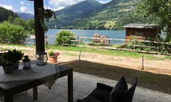 A beautiful view of a lake surrounded by mountains. In the foreground are a table and a chair on a terrace.