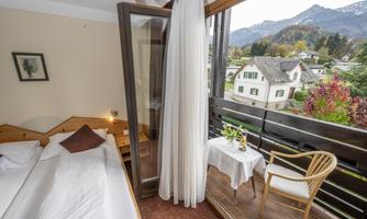 A cozy room with a balcony and a view of the mountains. A bed, a table, and a chair are visible, surrounded by a serene environment.