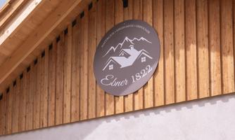 A traditional wooden house with a round sign displaying the name "Ebner 1822". The wooden windows and natural wood siding give the building a rustic charm.