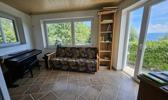 A cozy room with a sofa and a piano. Large windows offer a view of nature and plenty of light.