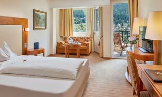 A cozy hotel room with a double bed, a table, and a balcony. Through the windows, there is a view of the mountains.