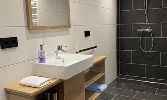 A modern bathroom with a sink on a wooden frame and a large mirror. The walls are brightly tiled, and a shower is visible in the back area.