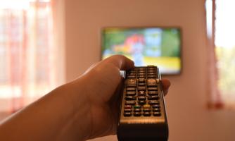 A hand holds a remote control in front of a television. The television displays a colorful image.
