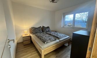 A cozy bedroom with a wooden bed and a bedside table. The room has a large window front that lets in light.