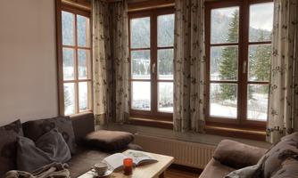A cozy living room with large windows that offer a view of snow-covered trees. A book lies on the table and a cup is beside it.