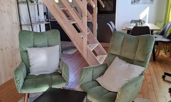 Two green armchairs are arranged around a small table in a bright room. In the background, a wooden staircase and a dining area are visible.