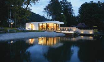 A modern house by the water with large windows and a terrace. The atmosphere is calm and illuminated in the evening.