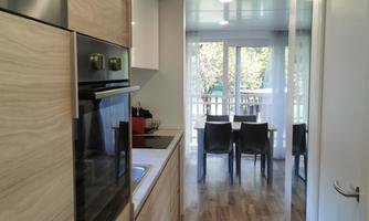 A modern kitchen with wooden cabinets and an oven. In the background, there is a table with chairs and plenty of natural light.
