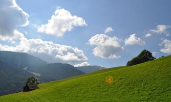 A green meadow with gentle hills and mountains in the background. The sky is blue with many white clouds.