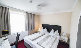 A bright bedroom with two single beds and white bedspreads. There is a window with curtains and modern furnishings.