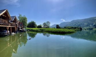 A quiet lake with wooden chalets and lush greenery. The mountains in the background and the clear sky create an idyllic atmosphere.